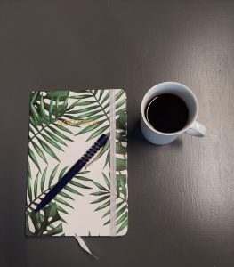 Kuvassa on kasvikuvioinen muistikirja ja sininen mustekynä, sekä kahvilla täytetty, valkea posliinikuppi.